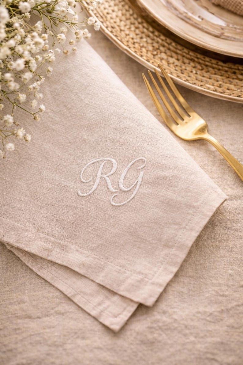 May include: Beige linen napkin with embroidered white initials "RG". A gold fork rests on the napkin, next to a woven plate. White flowers are in the upper left corner. The napkin is folded and has a textured appearance.