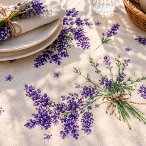 Può includere: Una tovaglia color crema con un motivo floreale di lavanda. Un mazzo di lavanda legato con spago poggia su un tovagliolo piegato sopra piatti bianchi. Sono visibili anche un cestino di vimini e un bicchiere d'acqua.
