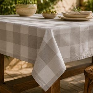 Op de afbeelding: Een rechthoekig tafelkleed met een groot ruitpatroon in afwisselende tinten zachtgrijs en wit. De stof valt over de randen van een houten tafel en vertoont een geweven textuur en overal strakke, gezoomde randen.
