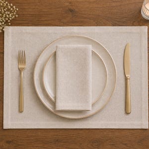 Op de afbeelding: Een gedekte tafel met een beige linnen placemat, twee gebroken witte borden, een gevouwen servet en goudkleurig bestek. Een brandende kaars en een klein bloemstuk staan op de achtergrond.