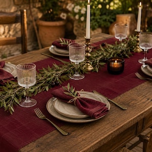 Peut inclure: Une table à manger en bois rustique avec un chemin de table en lin rouge foncé, des serviettes en tissu assorties nouées avec de la ficelle, des assiettes en céramique, des verres côtelés, des fourchettes dorées, des bougeoirs en laiton et une guirlande d'olivier.