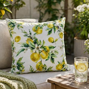 Könnte beinhalten: Ein dekoratives Kissen mit einem Aquarelldesign aus gelben Zitronen, grünen Blättern und weißen Blüten. Das Kissen liegt auf einem beigen Sofa mit einer grünen Decke. Ein Glas Wasser mit Zitronenscheiben steht auf einem Holztisch.