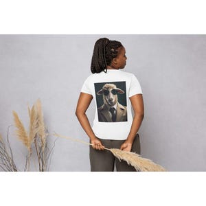 Op de afbeelding: Wit T-shirt met een afbeelding van een schaap met zonnebril en pak. De afbeelding staat op de achterkant van het shirt. Het model draagt een donkere broek en houdt een gedroogde plantenstengel vast. De achtergrond is effen grijs.