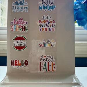 Puede incluir: Una colección de pegatinas de temporada con las frases «Hello Winter», «Hello Spring», «Hello Summer» y «Hello Fall». Las pegatinas presentan diseños coloridos, incluyendo copos de nieve, flores y una rodaja de sandía. Las pegatinas se muestran en un soporte blanco inclinado.