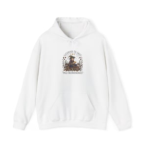 May include: White hooded sweatshirt with a front pocket and a drawstring. The graphic on the front features a witch with the text "LICENSED TO CAST" and "MILD INCONVENIENCES" in a circular design with botanical elements.