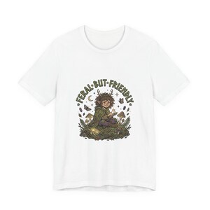 Puede incluir: Camiseta blanca con una ilustración de fantasía de una criatura del bosque, rodeada de setas y mariposas. El texto "FERAL BUT FRIENDLY" está arqueado sobre el personaje. El diseño utiliza tonos verdes, marrones y morados.