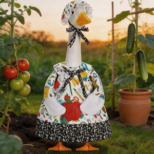 May include: A whimsical goose figurine dressed as a school teacher, wearing a patterned dress with school supplies, a bonnet, and a bow tie. The goose holds a red apple and stands in a garden with tomatoes and cucumbers.