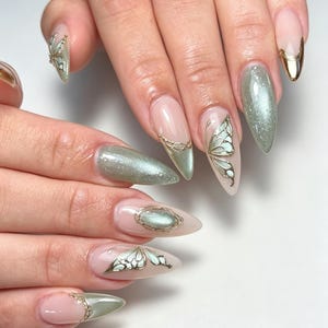 Puede incluir: Uñas en forma de almendra con base nude, detalles en oro metálico y esmalte verde salvia brillante. El diseño incluye motivos de alas de mariposa en oro, puntas francesas con borde dorado y un detalle central de piedra ovalada verde salvia.