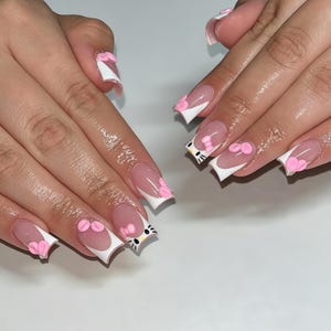 Puede incluir: Uñas postizas de forma cuadrada con estilo de manicura francesa, puntas blancas sobre una base color nude. El diseño incluye lazos rosas tridimensionales y un pequeño gráfico de la cara del personaje Hello Kitty en el dedo anular.