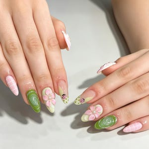 May include: Almond-shaped nails featuring a mix of designs including light pink grid patterns, solid sage green with white swirl accents, 3D pink floral embellishments with center crystals, and pale green tips with small painted ladybug motifs.