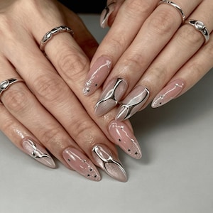 Op de afbeelding: Lange amandelvormige nagels met een nude basis, metallic zilveren vloeistofachtige accenten en kleine zwarte sterstickers. De vingers zijn versierd met drie zilveren ringen, waaronder een ring met textuur en twee gladde ringen met details.