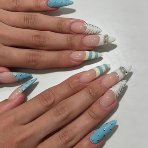 Op de afbeelding: Stiletto-vormige nagels met een mix van ontwerpen, waaronder lichtblauwe lak met zilveren studs, witte uiteinden met zwarte zebrastrepen, gele en blauwe horizontale strepen en witte uiteinden geaccentueerd met een band van kleine doorzichtige strass-steentjes.