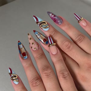 May include: Stiletto-shaped artificial nails featuring a variety of designs including red and blue plaid patterns, gold polka dots, vertical red stripes, and abstract red swirls. Several nails are accented with metallic gold rings and charms.