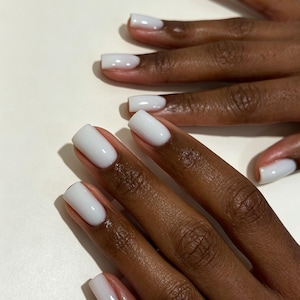May include: A set of fingernails with a glossy, opaque white finish. The nails are shaped in a square style with rounded edges, featuring a smooth, even application of polish that covers the entire nail bed from cuticle to tip.