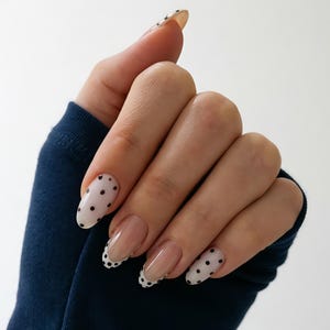 May include: A hand with long, oval-shaped nails. Some nails are painted with a white base and black polka dots. Other nails have a clear base with a white tip and black polka dots. The nails are a stylish accessory.