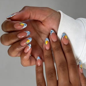 Puede incluir: Uñas en forma de almendra con una base nude y un diseño de manicura francesa. Las puntas están decoradas con un patrón floral azul y blanco e ilustraciones de limones amarillos. La manicura tiene un acabado brillante y forma redondeada.
