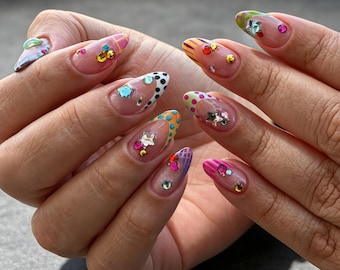 May include: Almond-shaped nails featuring a clear base with colorful French tips in patterns like polka dots, stripes, and grids. Each nail is decorated with various small, metallic, and gemstone-like embellishments in shapes including stars and circles.