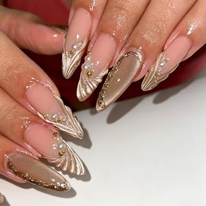 Peut inclure: Ongles longs en forme d'amande avec une base nude et des accents métalliques dorés texturés en forme d'éventail sur les pointes. Le design est orné de petites perles blanches et de minuscules billes dorées, avec un fini chatoyant effet œil-de-chat.