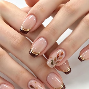 May include: Nails featuring a nude base with dark brown tips, accented by thin gold horizontal lines. Two nails display a delicate, hand-painted botanical leaf design in shades of brown and gold, adding a detailed element to the manicure.