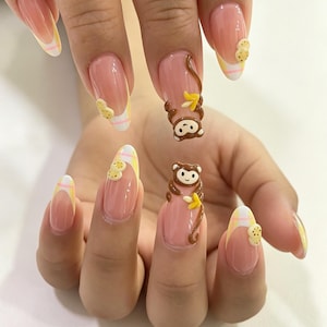 May include: Almond-shaped nails featuring a nude base with white and yellow plaid French tips. Two nails are adorned with 3D brown monkey figures holding yellow bananas, while other nails feature small 3D yellow cookie-like embellishments.