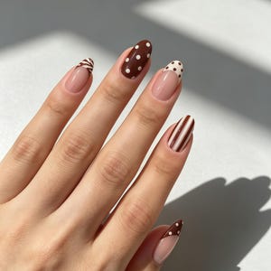 May include: A hand with almond-shaped nails featuring a variety of brown and white designs. The index finger has brown polish with white polka dots, the middle finger has a white tip with brown dots, the ring finger has brown and white vertical stripes, and the thumb and pinky feature brown tips with white polka dots.
