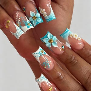 Puede incluir: Uñas postizas de forma cuadrada con una base de manicura francesa y patrones de ondas en azul claro y blanco. El diseño incluye flores tridimensionales en azul claro con centros dorados, pequeños detalles florales blancos, dijes de estrellas de mar doradas y diminutos diamantes de imitación azules.