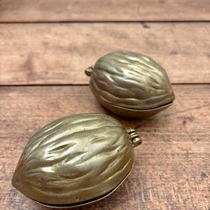 May include: Two brass objects shaped like walnuts with textured, ridged surfaces. Each piece features a hinged design, allowing the two halves of the shell to open. The metallic finish has a warm, golden tone with subtle variations in colour.