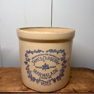 Könnte beinhalten: Ein zylindrischer, cremefarbener Keramik-Marmeladentopf mit dickem Rand. Auf der Vorderseite befindet sich ein blauer Aufdruck mit dem Text "Grand Medal for Marmalade James Carberry Trade Mark Marmalade London 1896 M&S" in einem floralen Rahmen.