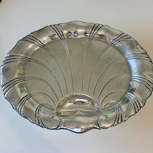 Puede incluir: Una fuente plateada con forma de concha. La fuente tiene un borde festoneado y un diseño en forma de abanico que irradia desde el centro. La fuente es ovalada y tiene una superficie lisa y reflectante.