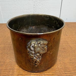 Könnte beinhalten: Ein Vintage-Weinkühler aus kupferfarbenem Metall. Der Eimer hat ein detailliertes Relief einer Weintraube auf der Vorderseite. Das Innere des Eimers ist dunkel, und die Außenseite hat eine warme, gealterte Patina. Er steht auf einer Holzoberfläche.