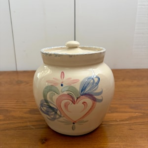 May include: A cream-coloured ceramic jar with a matching lid, featuring a hand-painted design of a central pink heart surrounded by blue and green swirling floral motifs. The rim of the lid has a subtle blue speckled pattern on the edge.