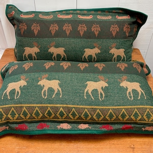 May include: Two rectangular throw pillows featuring a woven tapestry pattern in shades of forest green, tan, and red. The design includes repeating motifs of moose, maple leaves, canoes, and geometric diamond shapes with a solid green border.