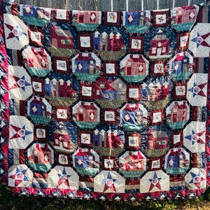 May include: A patchwork quilt featuring a repeating pattern of houses and barns in shades of red, blue, and green. The design includes star-shaped blocks with American flag motifs, interspersed with small square blocks and floral fabric accents.