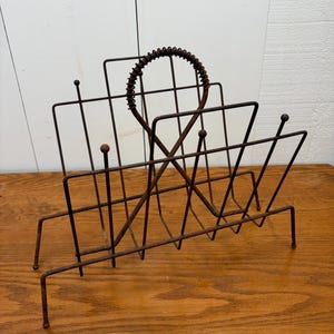 May include: A brown, rusted metal magazine rack featuring a wire grid construction with a central circular handle wrapped in coiled wire. The rack has four legs with small ball-shaped finials at the top of the vertical support rods.