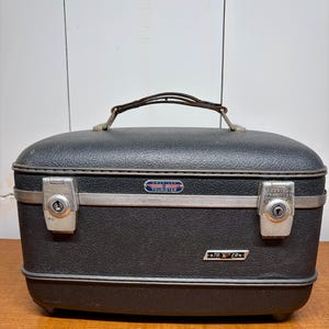 May include: A vintage black American Tourister vanity case with a textured exterior. It features a silver metal trim, two locking clasps, and a handle. The case has a rectangular shape with rounded edges.