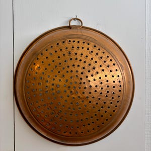 Puede incluir: Un colador redondo de cobre que presenta un patrón uniforme de pequeños orificios de drenaje circulares en toda su superficie. Este utensilio de cocina en forma de cuenco incluye una pequeña anilla metálica en el borde superior para colgarlo.