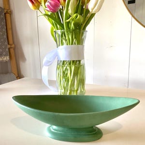 Könnte beinhalten: Eine grüne Keramik-Mittelpunktschale mit Sockel. Eine Glasvase mit weißem Band hält einen Strauß Tulpen in Rot-, Rosa- und Weißtönen. Die Anordnung steht auf einem weißen Tisch.