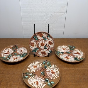 Könnte beinhalten: Set aus vier braunen Keramikplatten mit floralem Design. Jede Platte zeigt weiße, gänseblümchenartige Blumen mit braunen Zentren und grünen Blättern. Eine Platte wird auf einem schwarzen Ständer präsentiert.