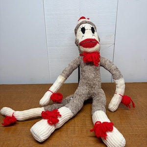 Puede incluir: Una muñeca de mono de calcetín hecha a mano con cuerpo marrón y blanco, boca roja y lazo rojo. La muñeca tiene piernas y brazos a rayas blancas y marrones, y pompones rojos unidos a las extremidades. El mono está sentado.