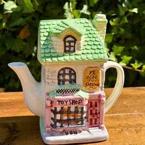 May include: A ceramic teapot shaped like a two-story building with a green shingled roof and a chimney. The facade features a pink storefront with a black barred window, a red door, and signs reading "TOY SHOP" and "YE OLDE TOY SHOPPE" in black text.