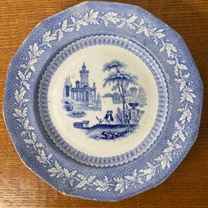 Peut inclure: Une assiette ancienne bleue et blanche avec une scène de paysage. Le bord est festonné et orné d'une bordure de feuilles. La scène centrale représente des bâtiments, des arbres et des personnages.