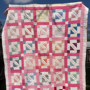 May include: A rectangular quilt featuring a grid of square blocks separated by bright pink sashing. Each block contains a diagonal strip of fabric with handwritten names in ink. The quilt is bordered by a wide pink fabric frame with white edges.