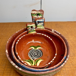 Könnte beinhalten: Ein Satz von drei ineinander verschachtelten, handbemalten Terrakotta-Schalen. Die Schalen sind rotbraun mit weißen, blauen, grünen und gelben Akzenten. Die kleinste Schale hat ein schwarz-weißes Spiraldesign mit grünen Blättern.