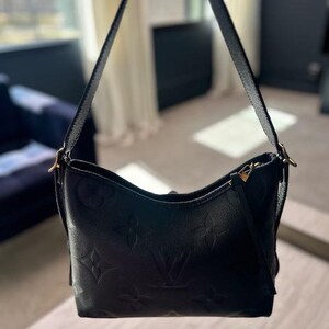May include: Black leather shoulder bag with embossed floral and logo pattern. The bag has a curved shape, a long shoulder strap, and gold-toned hardware. The bag is held up by a person.