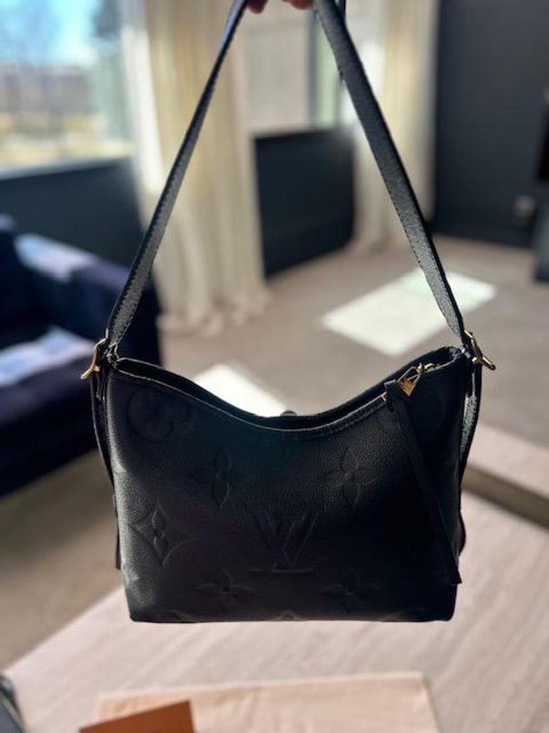 May include: Black leather shoulder bag with embossed floral and logo pattern. The bag has a curved shape, a long shoulder strap, and gold-toned hardware. The bag is held up by a person.