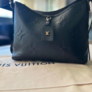 May include: Black leather shoulder bag with embossed floral pattern. Features a gold-tone tag with a logo. The bag has a black adjustable strap and is sitting on a beige surface with the brand name printed on it.