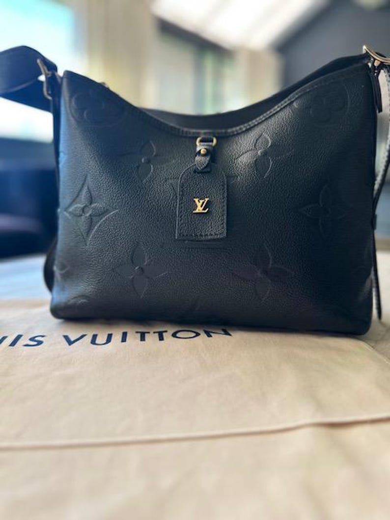 May include: Black leather shoulder bag with embossed floral pattern. Features a gold-tone tag with a logo. The bag has a black adjustable strap and is sitting on a beige surface with the brand name printed on it.
