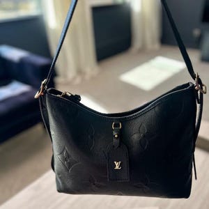 May include: Black leather shoulder bag with embossed floral pattern. Features a long strap, gold-tone hardware, and a small tag with a gold logo. The bag is displayed indoors with natural light.