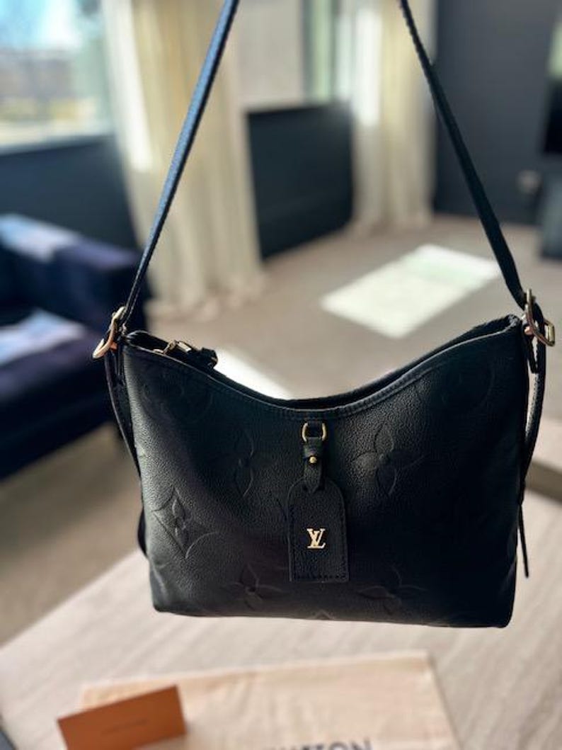 May include: Black leather shoulder bag with embossed floral pattern. Features a long strap, gold-tone hardware, and a small tag with a gold logo. The bag is displayed indoors with natural light.