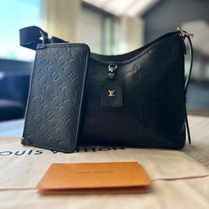 May include: Black leather shoulder bag and matching wallet with embossed monogram pattern. The bag features a top handle and a detachable strap. A small tag with a gold logo hangs from the bag. A small orange card sits in front of the bag.